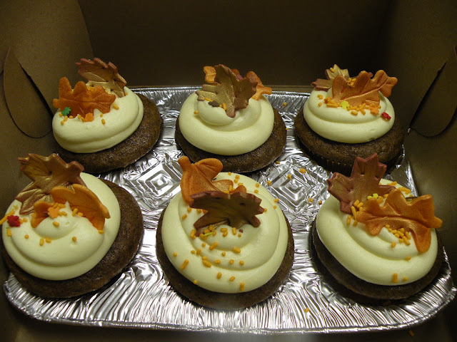 Six cupcakes with white frosting swirls topped with autumn-leaf decorations, arranged on a foil tray, warm mood